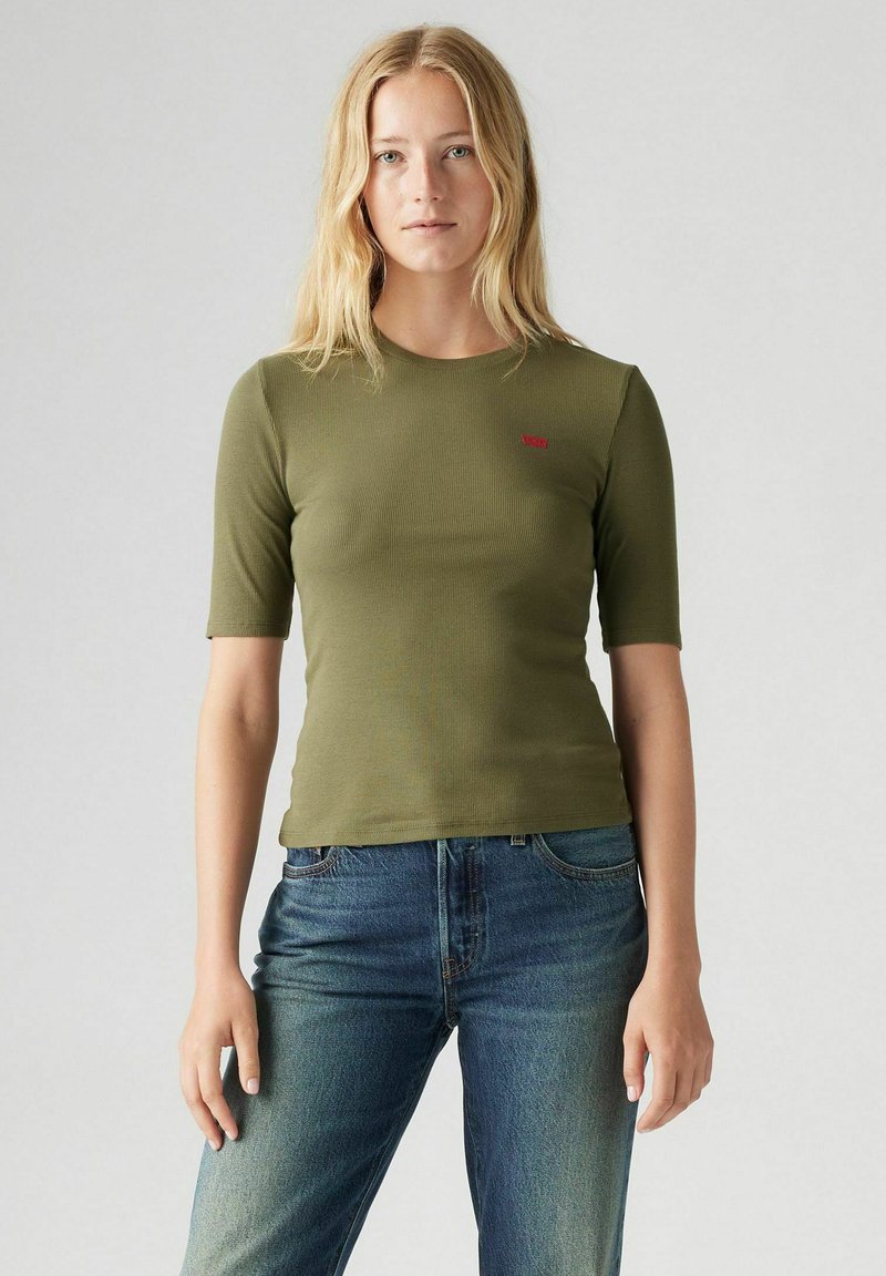 Grön ribbad kortärmad t-shirt med rund halsringning, prydd med en liten röd logotyp på vänster bröst, tillsammans med blå jeans.