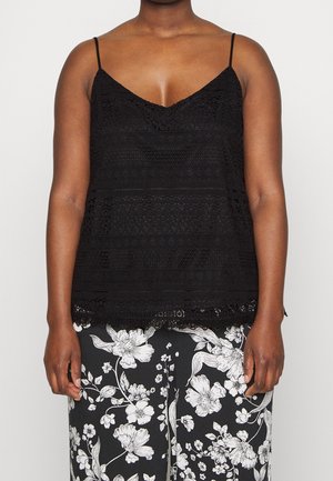 Femme portant un haut sans manches en dentelle noire avec des bretelles fines et un pantalon noir orné d'un grand motif floral blanc.