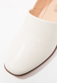 Chaussure slip-on en cuir blanc avec une texture lisse, un bout arrondi et une semelle marron subtile. Design minimaliste sans matériel visible.