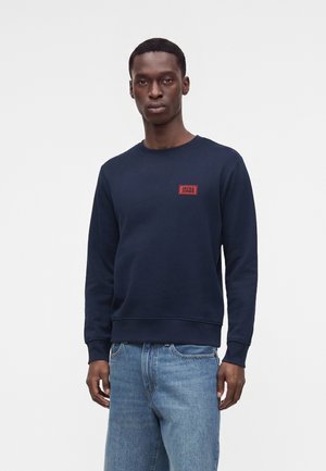 Mand iført en mørkeblå sweatshirt med en rød "Jack & Jones"-logo på brystet og lyseblå jeans mod en ensfarvet baggrund.