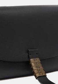 Borsa a tracolla in pelle nera con patta curvata. Presenta un accento in metallo bronzato con lettere incise e una texture liscia.