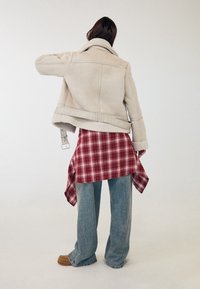 Personne debout de dos portant une veste en peau retournée beige, une chemise à carreaux rouge nouée à la taille, un jean bleu ample et des mocassins marron.