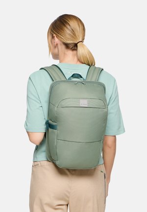 Persoon met blond paardenstaart die een lichtgroen shirt met korte mouwen en beige broek draagt, met een groene rechthoekige rugzak op de rug gericht.