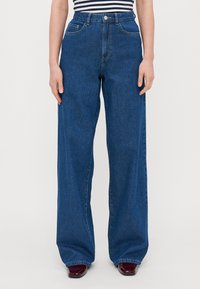 Jeans w szerokich nogawkach wykonane z ciemnoniebieskiego denimu. Posiadają wysoki stan, przednie kieszenie i zapięcie na guzik. Gładka tekstura z minimalnymi detalami szwów.