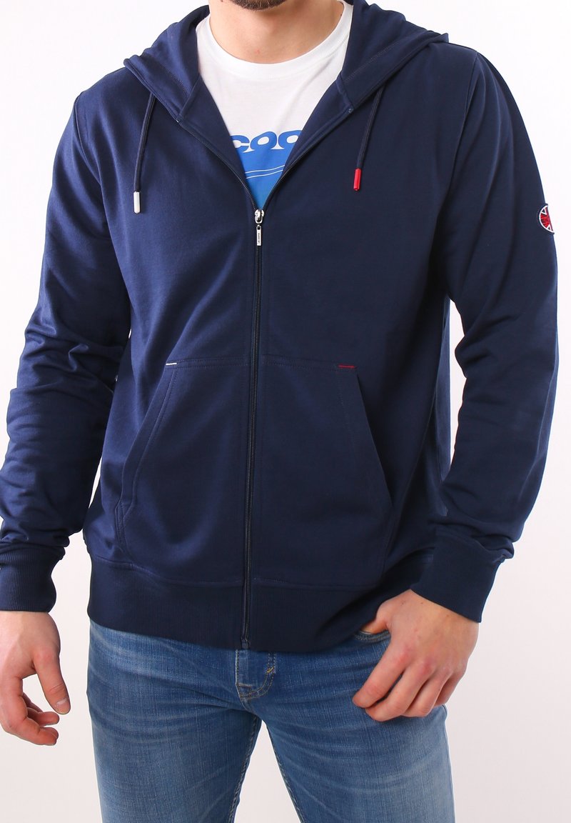 Granatowy hoodie na zamek wykonany z bawełny, z przednią kieszenią kangurką, ściągaczami przy rękawach oraz czerwonymi i białymi sznurkami do regulacji.