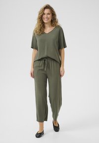 Combinaison ample de couleur vert olive avec un décolleté en V et des manches courtes. Présente une taille élastique à cordon de serrage et un design à jambes larges. Portée avec des ballerines noires.