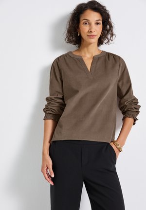 Femme aux cheveux bouclés portant une chemise marron à manches longues avec des poignets froncés et un pantalon noir, se tenant avec une main dans la poche contre un mur blanc.
