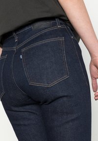 Jeans de ganga azul escura com costura visível, um patch de couro na cintura e dois bolsos traseiros. Textura suave, design ajustado.