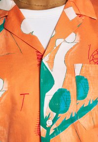 Koszula z krótkim rękawem na guziki, z pomarańczowym tłem i zielono-białymi abstrakcyjnymi wzorami, wykonana z lekkiego materiału o gładkiej fakturze.
