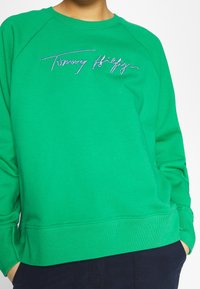 Sweatshirt vert avec des bords côtelés aux poignets et à l'ourlet. Présente un logo en strass "Tommy Hilfiger" sur le devant. Tissu doux, coupe décontractée.