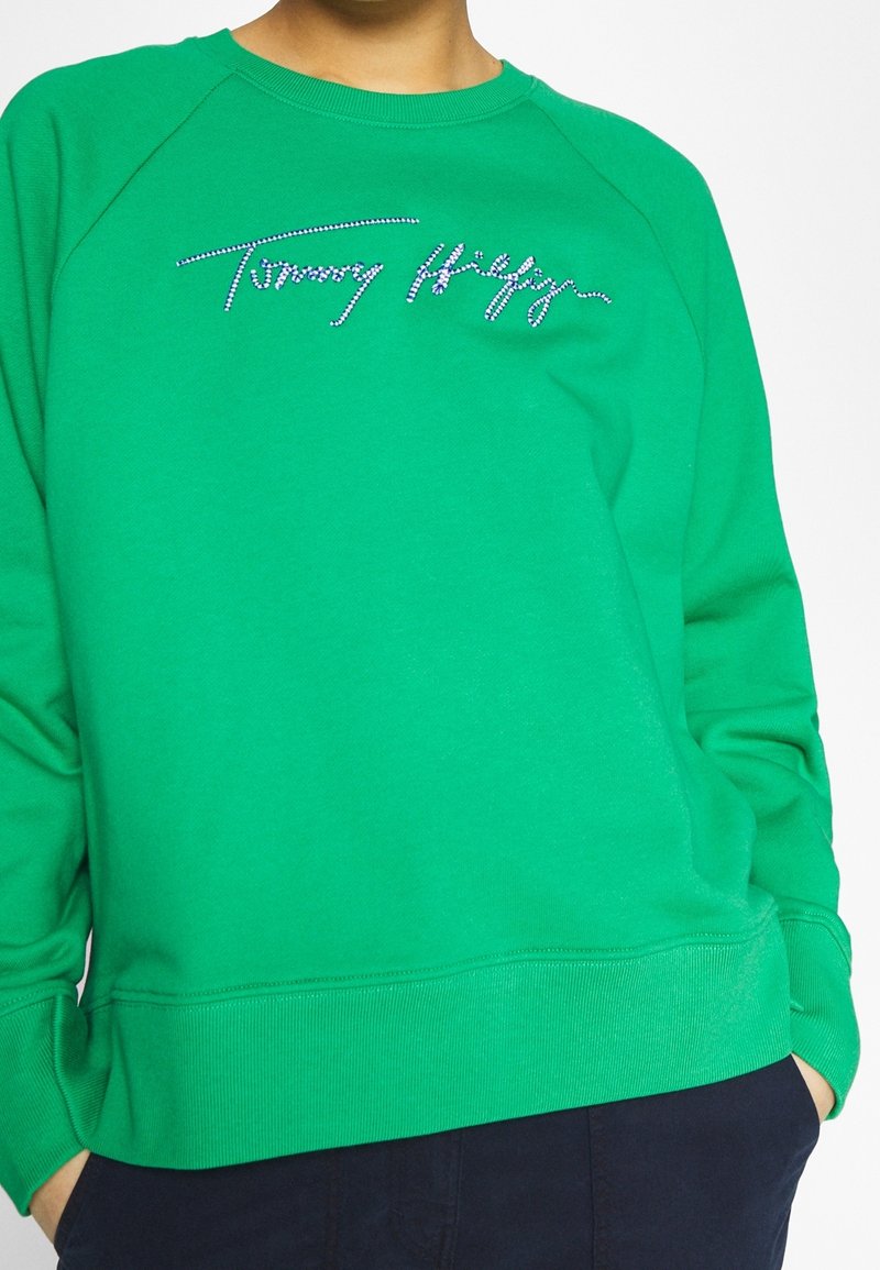 Sweatshirt vert avec des bords côtelés aux poignets et à l'ourlet. Présente un logo en strass "Tommy Hilfiger" sur le devant. Tissu doux, coupe décontractée.