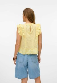Top giallo con volants e dettagli in pizzo, apertura a chiave sul retro e orlo svasato, abbinato a pantaloni corti in denim azzurro chiaro.