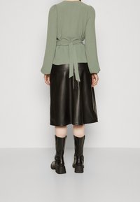 Haut vert texturé avec une taille nouée, associé à une jupe noir en cuir évasée. Des bottes noires jusqu'aux genoux complètent la tenue.