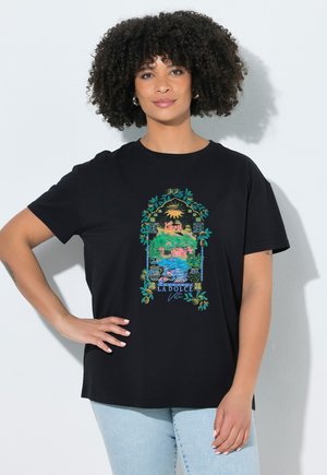 Kvinde med krøllet hår bærer sort T-shirt med farverig have- og vandscene indrammet af grønne vinranker, tekst "La Dolce Vita."