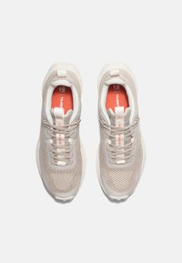 Zapatillas deportivas de color beige claro con parte superior de malla texturizada, detalles en crema y suelas blancas. Presentan cordones planos y una plantilla con branding en naranja.