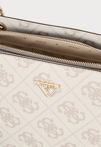 Guess handtas in beige synthetisch materiaal met geperst logo patroon, gouden beslag en ritssluiting. Gestructureerd oppervlak met een glanzende afwerking.