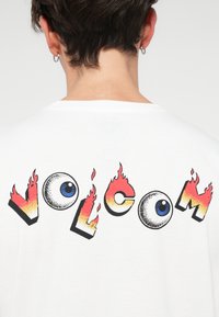 T-shirt in cotone bianco con una grafica colorata "VOLCOM", con lettere in fiamme nere e rosse, e un'illustrazione di un occhio al centro.
