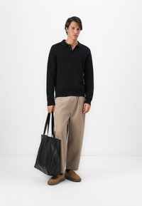 Jovem vestido com polo preto de manga longa, calças largas bege, sapatos castanhos, segurando uma grande bolsa de couro preta, em pé contra um fundo branco.