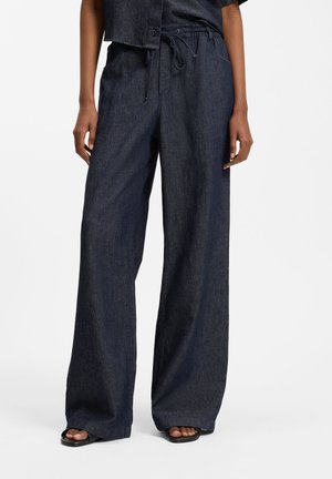 Pantalon large en denim foncé avec taille à cordon, porté par une personne debout, associé à des sandales noires à bouts ouverts.