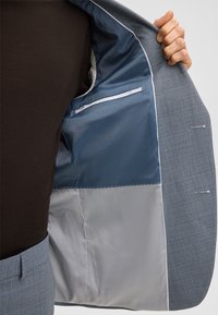 Grijs patroon colbert met een blauw gevoerde binnenkant. Voorzien van een zak met witte bies en een gladde textuur. Zichtbare arm die het colbert vasthoudt.