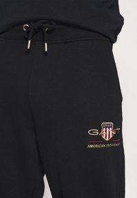 Zwarte sweatpants met een elastische tailleband, trekkoord en goud geborduurd logo met rode en witte accenten op de dij.