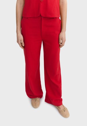 Osoba oblečená v voľných červených nohaviciach a zodpovedajúcom červenom bezrukávovom tričku, v béžových balerínach, stojaca pred jednoduchým pozadím.