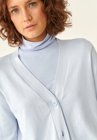 Hellblauer Cardigan mit V-Ausschnitt, einem Knopfverschluss, weichem Material und lockerer Passform, über einem hellblauen Rollkragenpullover getragen.