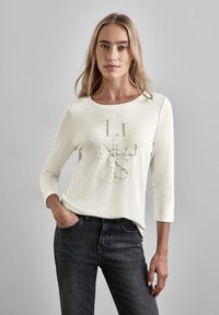 Lichtcrème lange mouw shirt met een textuur grafisch ontwerp waarin de woorden "LINES" en "Casual" in subtiele tinten zijn weergegeven, gecombineerd met zwarte jeans.