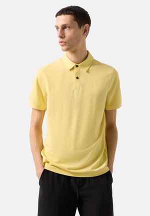 Young man with short dark hair wearing a yellow short-sleeve polo shirt and black pants, hands in pockets, looking left.