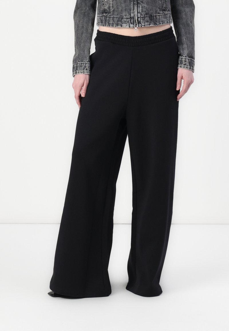 Personne portant un pantalon noir à jambes larges avec une taille élastique et une veste en jean noire délavée courte sur un fond blanc uni.
