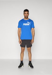 Zils īsām piedurknēm T-kreklis ar baltu "PUMA" logotipu, apvienots ar melniem sporta šortiem un melniem sporta apaviem. Vienkārša fona.