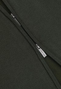 Ciemnozielony sweter z teksturowanym splotem, z czarnym zamkiem błyskawicznym na środku i małą metką przyczepioną do zamka.