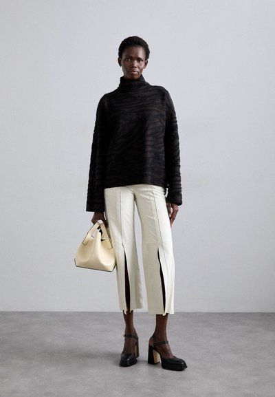 Suéter negro texturizado con cuello alto, pantalones blancos de pierna partida y un pequeño bolso crema. La modelo lleva zapatos negros de tacón cuadrado.