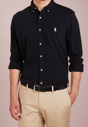 Mężczyzna ubrany w czarną, rozpinaną koszulę z długim rękawem, z podwiniętymi rękawami, oraz beżowe spodnie z czarnym paskiem, stojący na jednolitym tle.