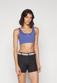 Sujetador deportivo morado con un logo, combinado con pantalones cortos negros que tienen una cintura de marca blanca. Tejido suave, diseño ajustado.