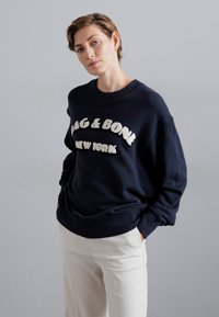 Granatowy sweter z napisem "RAG & BONE NEW YORK" w puszystym, białym kolorze, okrągły dekolt, luźny krój i ściągacze przy rękawach. Spodnie mają kremowy kolor.