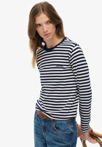 Kvinde med langt hår iført en navy- og hvidstribet langærmet skjorte og blå jeans, som holder en træstol foran en hvid baggrund.