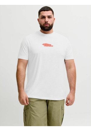 Homme avec barbe portant un t-shirt blanc à motif graphique et un short cargo olive, debout devant un fond clair uni.