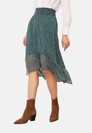 Femme portant une jupe midi verte à motifs taille haute avec un ourlet volanté et des bottines en daim marron, associée à une chemise blanche à manches longues.