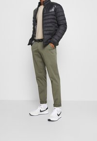 Schwarze Pufferjacke mit glänzender Oberfläche, beiger Pullover, olivgrüne Hose, schwarzer Gürtel und weiße Sneakers mit schwarzen Akzenten.