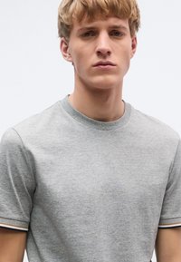 Szürke rövid ujjú póló puha anyagból készült. Kerek nyakkivágással és kontrasztos csíkos díszítéssel a ujjvégeken.