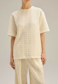 Kremowy, luźny top o teksturowanym, falistym wzorze. Materiał jest miękki, a krótkie rękawy nadają swobodny wygląd.