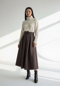 Kremowy sweter z dzianiny z golfem w połączeniu z brązową plisowaną spódnicą z wysokim stanem i czarnymi botkami, podkreślający gładkie tekstury i krągłości.