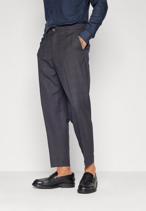 Mies, jolla on päällään tummat ruudulliset housut ja mustat nahkakengät ilman nauhoja, toinen käsi taskussa, seisoo yksinkertaisen vaalean taustan edessä.