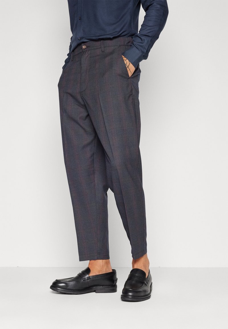 Homme portant un pantalon à carreaux foncés et des mocassins en cuir noir, une main dans la poche, debout devant un fond clair uni.