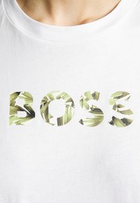 T-shirt bianca in cotone con la parola "BOSS" in lettere grasse, riempita da un motivo di foglie di palma verde. Maniche corte, scollatura rotonda.