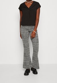 Top nero a maniche corte con scollo a V, abbinato a pantaloni svasati grigi e neri con motivo. La modella indossa scarpe nere chunky e gioielli semplici.