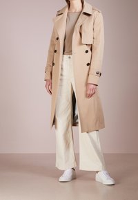 Impermeabile beige con design doppiopetto, grandi bottoni neri e spalline, abbinato a pantaloni bianchi a gamba larga e sneakers bianche.