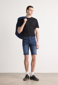 T-shirt nera con logo, pantaloni corti di denim blu, calzini bianchi e scarpe nere. Il modello tiene una giacca di denim blu sulla spalla.