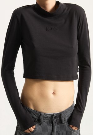 Personne portant un haut court noir à manches longues et un pantalon taille haute gris à carreaux, les mains dans les poches, dévoilant le ventre.